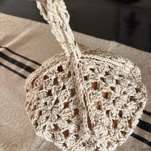 Op de afbeelding: Een cr&egrave;mekleurige gehaakte tas met een trekkoordsluiting en een lusvormige handgreep. De tas heeft een bloemenpatroon en spikkels groen en oranje. De tas ligt op een beige en zwart gestreept oppervlak.