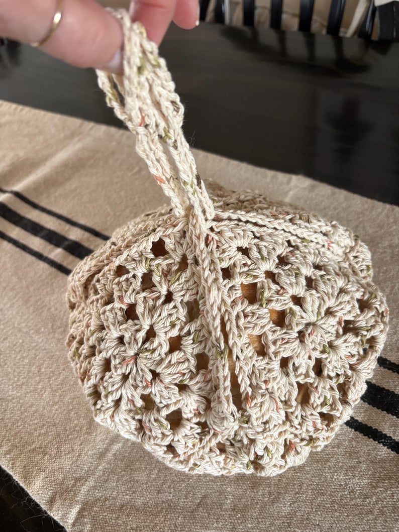 Op de afbeelding: Een cr&egrave;mekleurige gehaakte tas met een trekkoordsluiting en een lusvormige handgreep. De tas heeft een bloemenpatroon en spikkels groen en oranje. De tas ligt op een beige en zwart gestreept oppervlak.