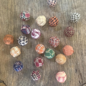 Pu&ograve; includere: Una collezione di venti piccole perline sferiche rivestite in tessuto, ognuna caratterizzata da un motivo a quadri unico in vari colori tra cui rosso, blu, verde, arancione, marrone, beige e bianco, sparse su una superficie di legno.