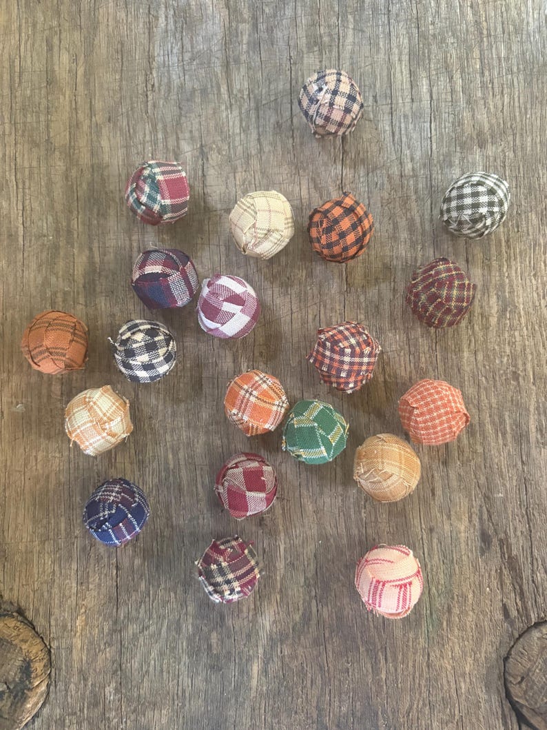 Pu&ograve; includere: Una collezione di venti piccole perline sferiche rivestite in tessuto, ognuna caratterizzata da un motivo a quadri unico in vari colori tra cui rosso, blu, verde, arancione, marrone, beige e bianco, sparse su una superficie di legno.