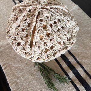 Op de afbeelding: Een gehaakte broodhoes in cr&egrave;me en gespikkeld garen, bovenaan vastgebonden. De hoes ligt op een beige linnen loper met zwarte strepen. Twee takjes rozemarijn zijn bij de broodhoes geplaatst.
