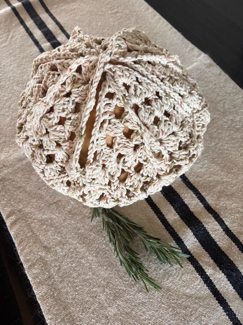 Op de afbeelding: Een gehaakte broodhoes in cr&egrave;me en gespikkeld garen, bovenaan vastgebonden. De hoes ligt op een beige linnen loper met zwarte strepen. Twee takjes rozemarijn zijn bij de broodhoes geplaatst.