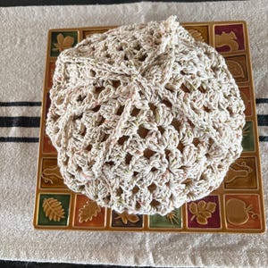 May include: A crocheted hat in a neutral color with flecks of green and pink. The hat is sitting on a decorative square tile with autumn leaf designs in various colors.