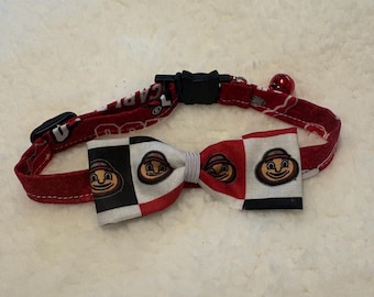 Op de afbeelding: Een rode stoffen halsband voor huisdieren met een zwarte plastic gesp en een klein rood belletje. Aan de halsband zit een strikje met een rood, wit en zwart ruitpatroon, versierd met herhalende cartoongezichten die hoeden dragen.