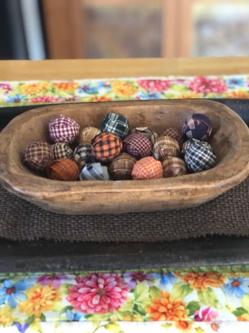 Pu&ograve; includere: Una ciotola in legno poco profonda di forma ovale riempita con una collezione di piccole sfere rivestite in tessuto. Le sfere presentano vari motivi a quadri e scozzesi in colori tra cui arancione, marrone, blu, verde e bianco.