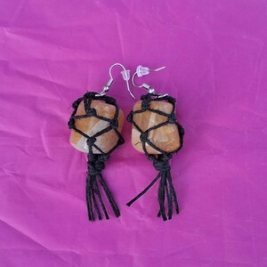May include: A pair of handmade earrings featuring orange-toned stones wrapped in black macrame. The earrings have silver-colored ear wires and a tassel design at the bottom. The background is a solid, bright pink color.