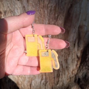 May include: A pair of yellow and orange gas pump earrings. The earrings are made of a translucent material and hang from silver-colored hooks. The gas pump design includes a nozzle and a rectangular display.