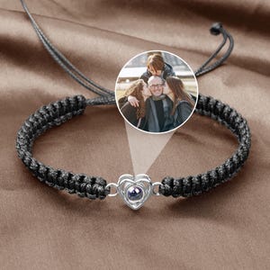 May include: Black braided bracelet with a silver heart-shaped charm. The charm has a small, dark center and is connected to the bracelet with silver links. A circular image of a family is projected above the bracelet.