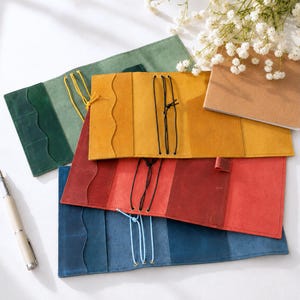 May include: A collection of four rectangular leather notebook covers in green, yellow, red, and blue, each featuring internal pockets with wavy edges and elastic cord closures. A white fountain pen is positioned to the left of the covers.