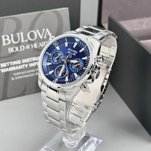 May include: A silver-tone stainless steel Bulova Marine Star chronograph watch with a deep blue dial, three sub-dials, luminous hands, and a date window. The watch features a polished link bracelet and a rotating bezel with numerical markings.