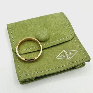 May include: A plain gold band ring resting on a light green suede pouch. The pouch features a flap with a button closure and a white embroidered logo on the bottom right corner that displays the letters VCA with a central pillar icon.