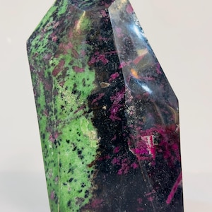 Op de afbeelding: Een gepolijste, gefacetteerde edelsteen met een mix van kleuren. De steen heeft een basis van zwart en donkergroen, met levendige magenta en roze insluitsels. De bovenkant is schuin en de algemene vorm is een hoog, rechthoekig prisma.