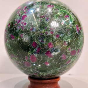 Könnte beinhalten: Eine polierte Edelstein-Kugel, überwiegend grün mit leuchtend roten und blauen Einschlüssen. Die Kugel ist auf einem kleinen, runden Holzsockel montiert. Die Oberfläche des Edelsteins reflektiert das Licht und hebt seine vielfältigen Texturen und Farben hervor.