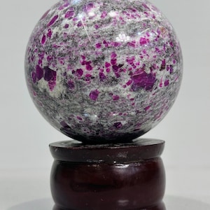 May include: A polished sphere of ruby in granite, resting on a dark wooden stand. The sphere is primarily gray with vibrant magenta ruby inclusions. The stand is a rich, dark brown, adding a touch of elegance to the display. A decorative item.