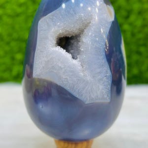 Puede incluir: Un huevo de ágata pulida, azul grisáceo, con una cavidad de geoda llena de formaciones cristalinas blancas. El huevo está montado sobre un pequeño soporte de madera torneada, mostrando la belleza natural de la piedra. El fondo es verde borroso.