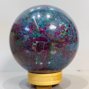Puede incluir: Un espécimen mineral esférico pulido con una superficie moteada de azul profundo, magenta vibrante y toques de verde. La esfera descansa sobre un pequeño soporte cilíndrico de madera clara, mostrando una textura suave y reflectante.