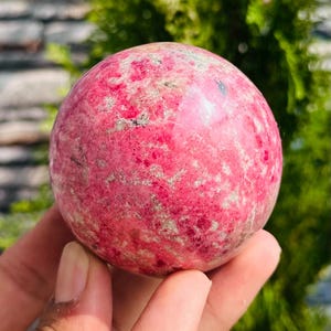 Peut inclure: Une pierre précieuse sphérique polie dans les tons de rose et de rouge, avec de subtiles veines grises et blanches. L'orbite est tenue dans une main, avec un arrière-plan vert flou.