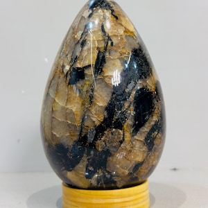 Puede incluir: Una escultura de piedra pulida en forma de huevo con un patrón complejo de inclusiones minerales translúcidas en tonos beige, ocre y negro opaco. El objeto liso y brillante se exhibe verticalmente sobre una base de madera clara.