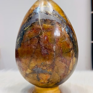 Peut inclure: Une sculpture en pierre polie en forme d'œuf présentant un motif complexe de veines ambrées chaudes, rouille profonde et gris anthracite foncé. La surface est lisse et brillante, révélant des inclusions minérales et des fissures naturelles.