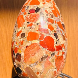 Op de afbeelding: Een eivormige stenen sculptuur met een mozaïek oppervlak. Het heeft rode, oranje, zwarte en beige fragmenten. De sculptuur staat op een zwarte metalen standaard, die de gevarieerde texturen en kleuren benadrukt. Hoogte ongeveer 10 cm.