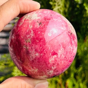 Peut inclure: Une pierre précieuse sphérique polie, dans des tons de rose foncé et de rouge, avec de subtiles variations de couleur et de texture. La sphère est tenue dans une main, avec un arrière-plan vert flou.