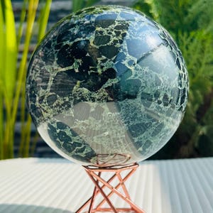 May include: A large, polished sphere with a mottled pattern of dark green and black, displayed on a geometric copper-coloured metal stand. The sphere reflects light, and the stand has a hexagonal base.