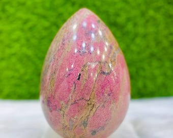 May include: A polished, teardrop-shaped gemstone in shades of pink and tan, mounted on a small wooden base. The stone has a smooth, reflective surface with natural veining. The background is a vibrant green.