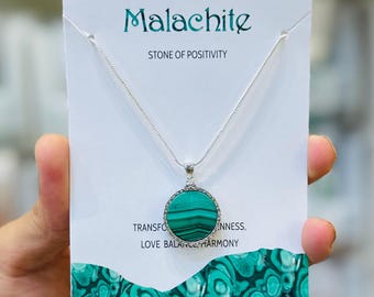 Pode incluir: Um colar de prata com um pingente redondo de malaquite. O pingente é verde com listras verde-escuras e é emoldurado em prata. O colar é exibido em um cartão branco com a palavra "Malaquite" e a frase "STONE OF POSITIVITY".