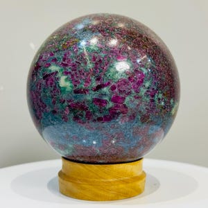 May include: A polished spherical stone featuring a mottled pattern of deep magenta, dark green, and blue-grey mineral inclusions. The sphere rests securely on a smooth, light-colored wooden circular stand with a visible natural grain.