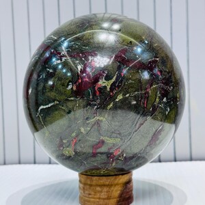 May include: A polished, spherical gemstone with a mix of green, red, and gold veining. The orb is displayed on a small, round wooden stand. The stone's surface is glossy and reflective, showcasing its natural patterns.