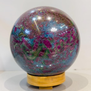 May include: A polished spherical mineral specimen featuring a vibrant mix of deep magenta, blue, and green crystalline inclusions throughout its surface. The sphere is supported by a simple, light-colored circular wooden stand for display.