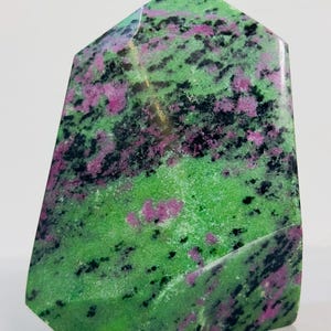 Peut inclure: Cristal de zoïsite poli avec inclusions de rubis. La pierre présente une base verte vibrante, parsemée de noir et de taches roses et violettes. Le cristal est taillé en forme géométrique avec une base plate et des côtés inclinés.
