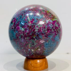 Pode incluir: Uma esfera mineral polida apresentando uma mistura vibrante de cristais de rubi vermelho magenta profundo incrustados em uma matriz de cianita azul e fuchsite verde, apoiada sobre um pequeno suporte circular de madeira castanho-claro.