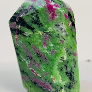 May include: A polished, faceted gemstone with a vibrant green base color, accented with streaks of deep pink and black. The stone has a smooth, glossy surface and a unique, irregular shape. The gemstone is a decorative item.