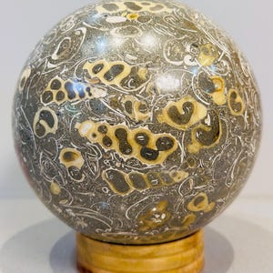 May include: A decorative sphere with a unique pattern of gray, cream, and tan markings. The sphere is displayed on a small, round, wooden stand. The stone has a polished finish, showcasing the natural variations in color and texture.