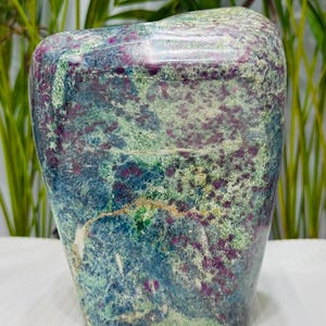 Peut inclure: Une pierre rectangulaire polie avec un mélange de couleurs. La pierre présente une base de teintes bleues et vertes, avec des taches de violet profond et une texture tachetée. La pierre est lisse et a un dessus arrondi.