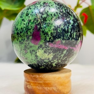 Op de afbeelding: Een gepolijste bolvormige steen met een gevlekt patroon van diep bosgroen, zwart en levendige magenta insluitingen. De bol rust stevig op een cilindervormige, lichtbruine houten standaard met zichtbare natuurlijke nerven en textuur.
