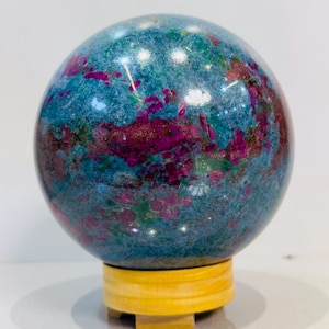 Può includere: Un esemplare minerale sferico lucidato caratterizzato da un vibrante colore di base blu e verde acqua con prominenti inclusioni di macchie cristalline color magenta intenso e fucsia su tutta la superficie, poggiato su un supporto circolare in legno.