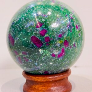 Può includere: Una sfera di pietra preziosa lucidata, con un vibrante colore di base verde, macchiata di inclusioni magenta intenso. La sfera poggia su un piccolo supporto rotondo di legno, mettendo in risalto la bellezza naturale della pietra.