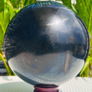 Könnte beinhalten: Eine polierte, dunkelgraue Kugel, möglicherweise ein Edelstein oder ein dekoratives Objekt, steht auf einem tiefroten Holzsockel. Die Kugel reflektiert das Licht. Im Hintergrund ist grünes Laub zu sehen.