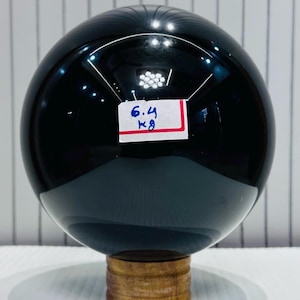 Op de afbeelding: Een grote, gepolijste zwarte bol, mogelijk een decoratief object, staat op een kleine houten standaard. Een wit label met de tekst "6,4 kg" is bevestigd. Het oppervlak reflecteert licht. De bol weegt 6,4 kilogram.