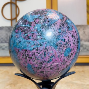 Può includere: Una pietra sferica lucida caratterizzata da un mix vibrante di inclusioni minerali color magenta, blu brillante e nero. La sfera poggia saldamente su un supporto in metallo nero a tre punte, mostrando una superficie liscia e riflettente.