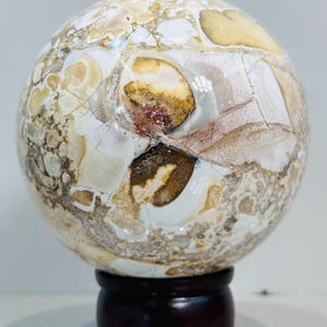 Puede incluir: Una esfera decorativa con un patrón único en colores beige, marrón y blanco. La esfera está montada sobre un soporte de madera marrón oscuro. La superficie de la esfera tiene un aspecto agrietado, similar a un mosaico, con áreas de color marrón rojizo y beige.