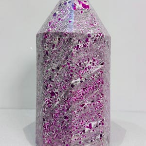 Puede incluir: Una punta de cristal hexagonal con un acabado pulido. La piedra presenta una mezcla de gris y blanco con vibrantes inclusiones magenta y moradas. El cristal tiene una parte superior puntiaguda y una base plana.