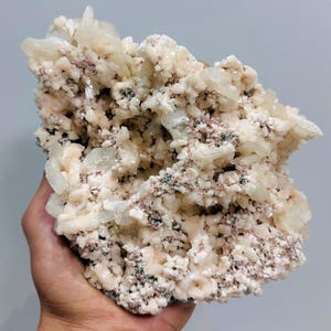 May include: A large, natural mineral specimen featuring clusters of off-white, crystalline formations. The mineral has a rough, textured surface with hints of pink and green throughout. The overall shape is irregular and organic, showcasing the raw beauty of the stone.