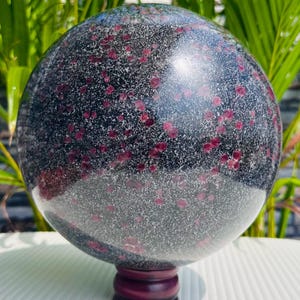 Può includere: Una sfera lucida di granito nero macchiata di cristalli di granato rosso, montata su un supporto in legno viola scuro. La superficie lucida della sfera riflette la luce. L'oggetto decorativo è adatto all'esposizione. Diametro di circa 15 cm.