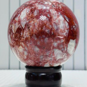 Peut inclure: Une sphère décorative polie, présentant des motifs rouges et blancs. La sphère est posée sur un socle en bois brun foncé. Cet objet décoratif est idéal pour la décoration intérieure.
