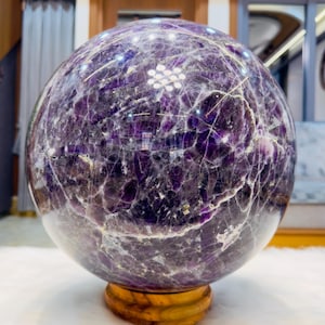 Könnte beinhalten: Eine große, kugelförmige Amethystkristallkugel mit tiefvioletten Farbtönen und komplexen weißen sowie durchscheinenden Adern auf der polierten Oberfläche. Der Stein ist auf einem runden, hellbraunen Holzsockel befestigt.