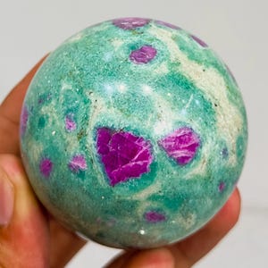 May include: A polished gemstone sphere with a teal base color and vibrant magenta inclusions. The sphere's surface has a mottled appearance, with the magenta inclusions varying in size and shape, creating a unique pattern. The sphere is held in a hand.