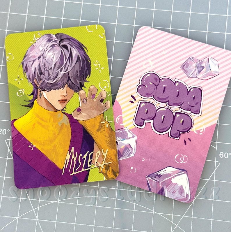 K&ouml;nnte beinhalten: Zwei rechteckige Sammelkarten. Die linke Karte zeigt eine Anime-Figur mit violettem Haar, gelbem Rollkragenpullover und violetter Weste, mit dem Wort MYSTERY in Gelb. Die rechte Karte ist rosa mit dem Text SODA POP und Eisw&uuml;rfeln.